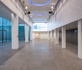 Eingangsbereich Foyer der ConferenceArea im Haus der Bayerischen Wirtschaft. Das Bild zeigt eine große Halle mit weißen Säulen und blauer Ambience-Beleuchtung. Am Ende der Halle ist eine große LED-Wand, mit Logo des Hauses.