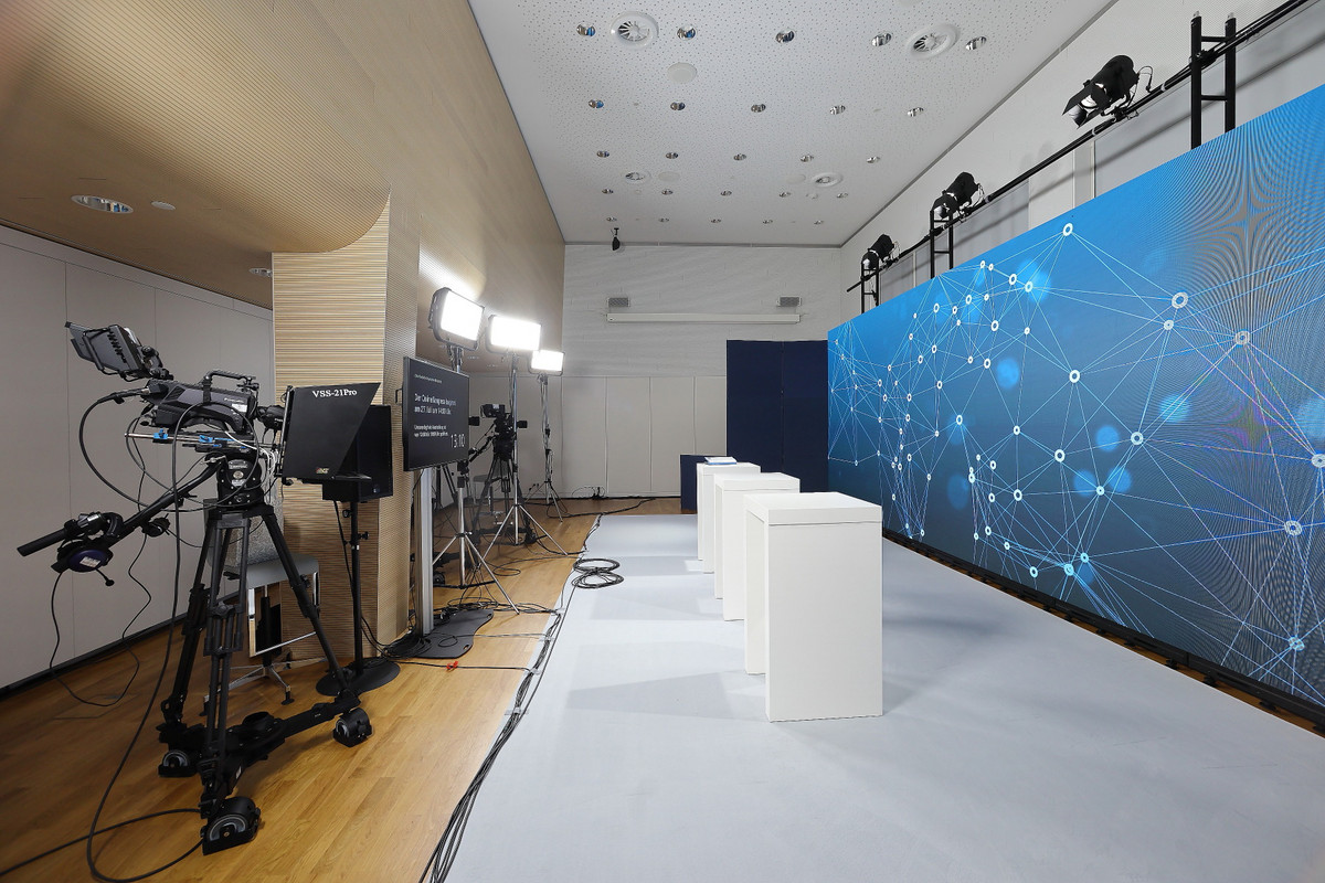 Modernes Streaming Setting mit LED Rückwand. Aufbau einer Pressekonferenz im Münchner Saal im Haus der Bayerischen Wirtschaft. Auf der linken Seite stehen professionelle Studiokameras. An der rechten Wand ist über die gesamte Länge des Raums eine LED-Wand aufgebaut. Die Wand ist mit einer futuristischen Grafik in Blautönen bespielt. Vor der LED-Wand stehen weiße Quader, die als Stehpulte für die Redner dienen.