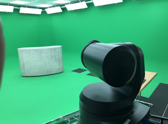 Blick von der Regie des GreenScreenStudios in den Aufnahmeraum. Im Vordergrund sieht man die PTZ-Kamera. Im Aufnahmeraum, dess Wändegrün gestrichen sind, steht ein Tresen in Betonoptik.