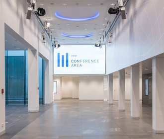 Eingangsbereich Foyer der ConferenceArea im Haus der Bayerischen Wirtschaft. Das Bild zeigt eine große Halle mit weißen Säulen und blauer Ambience-Beleuchtung. Am Ende der Halle ist eine große LED-Wand, mit Logo des Hauses.