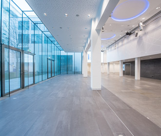 lichtdurchflutetes Foyer mit deckenhohen Glasscheiben im Erdgeschoß im Haus der Bayerischen Wirtschaft. Das Bild zeigt den großen Eingangsbereich mit weißen Säulen und blauer Ringbeleuchtung